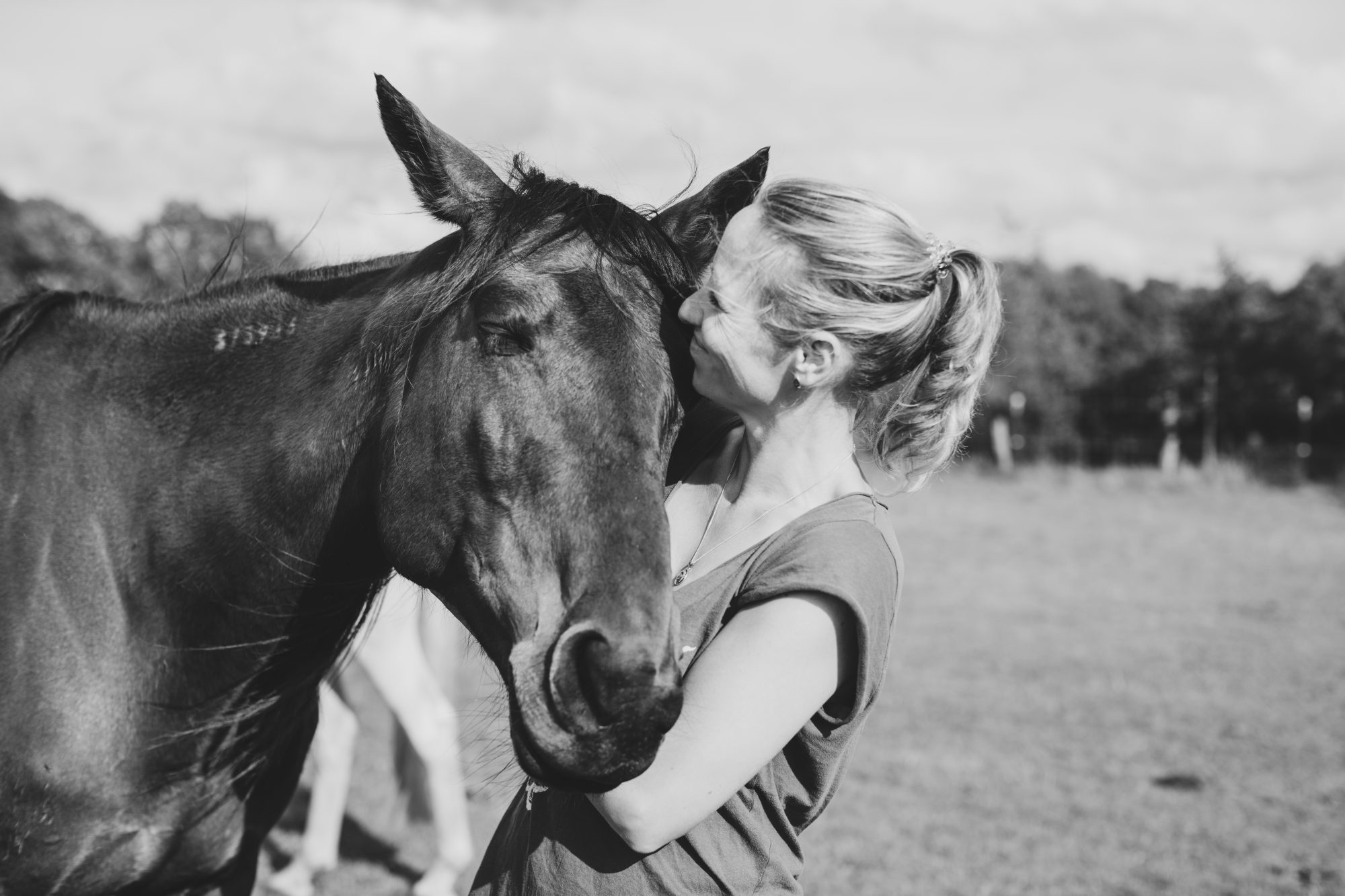 Schwarzweißaufnahme einer Frau in engem Kontakt mit einem Pferd auf einer Wiese.