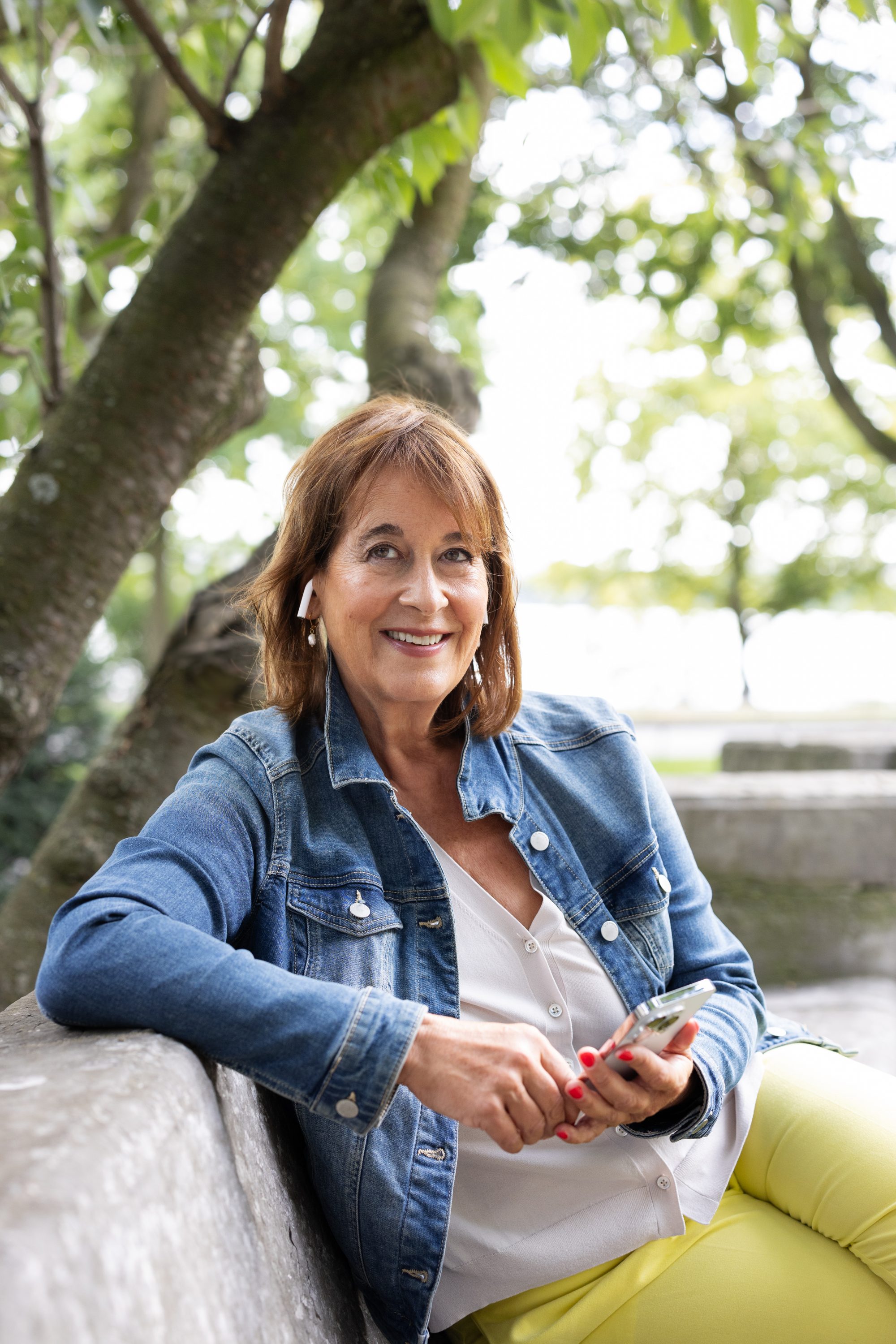 Lächelnde Frau in einer Jeansjacke vor Bäumen im Freien.