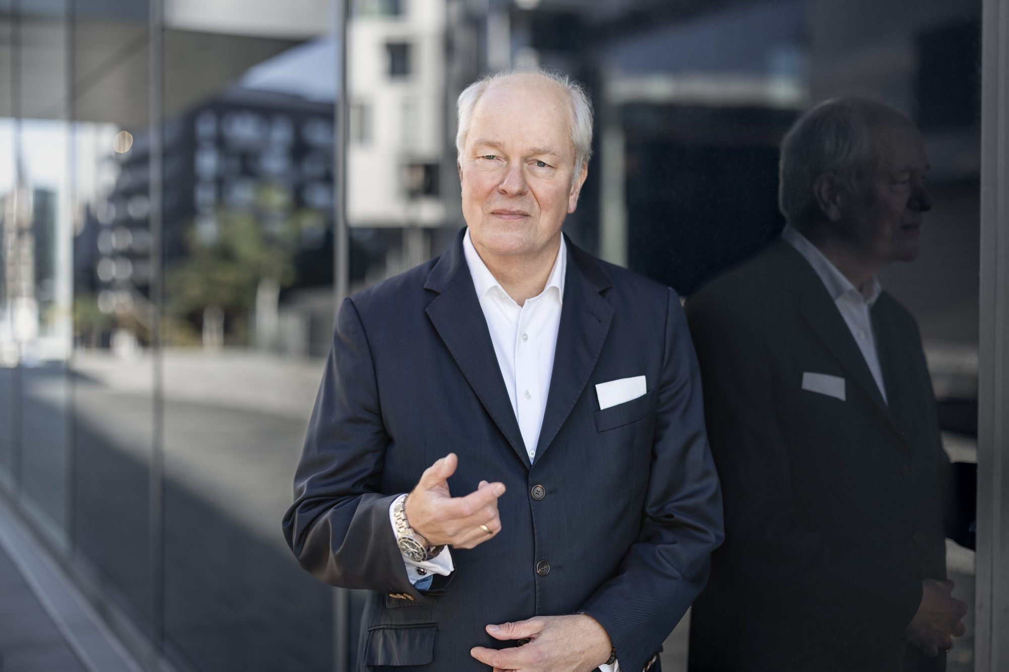 Henning von Gersdorff gestikuliert vor einer dunklen, spiegelnden Glasfassade.