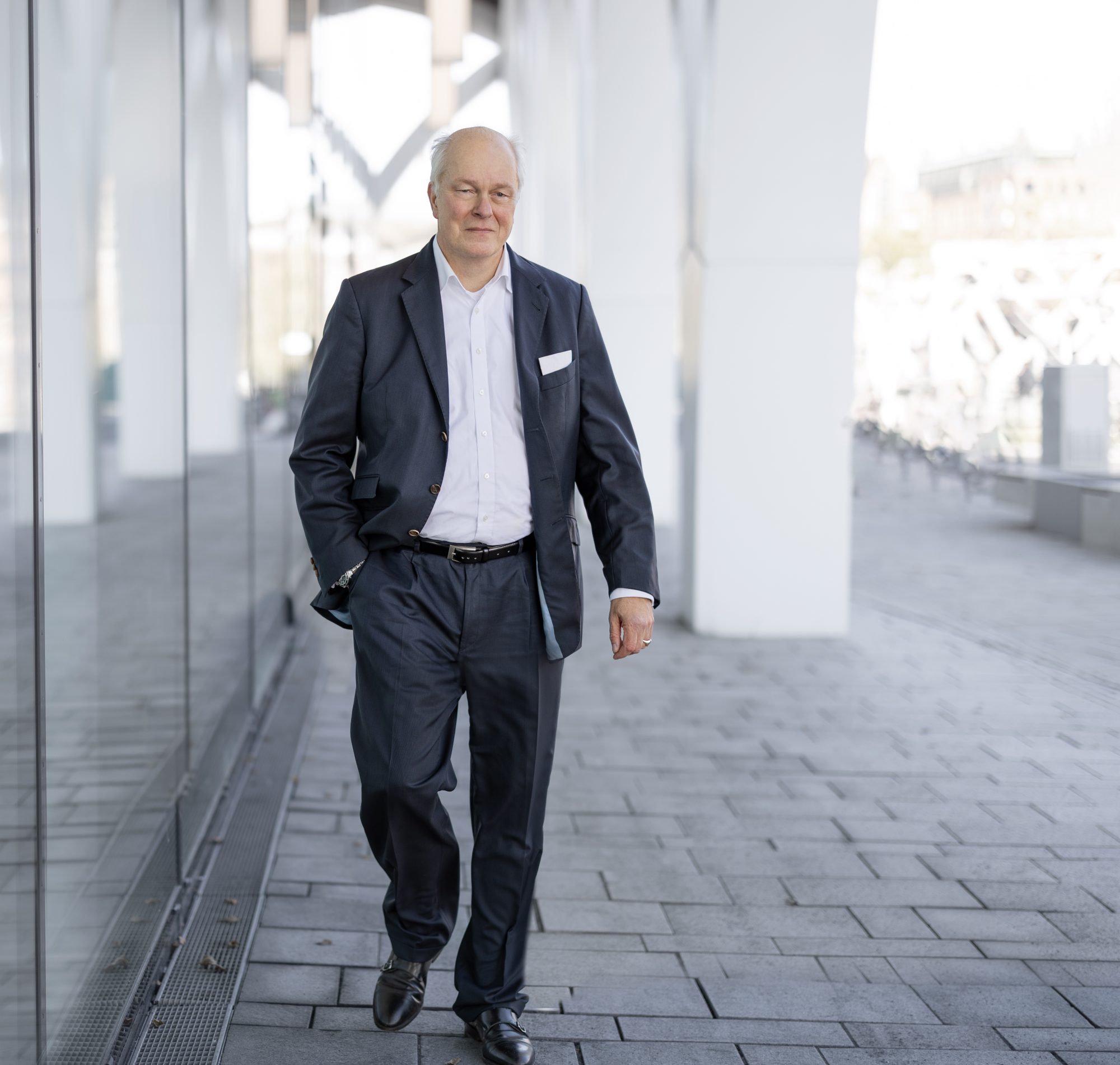 Henning von Gersdorff geht im Anzug entlang einer hellen Gebäudefassade.