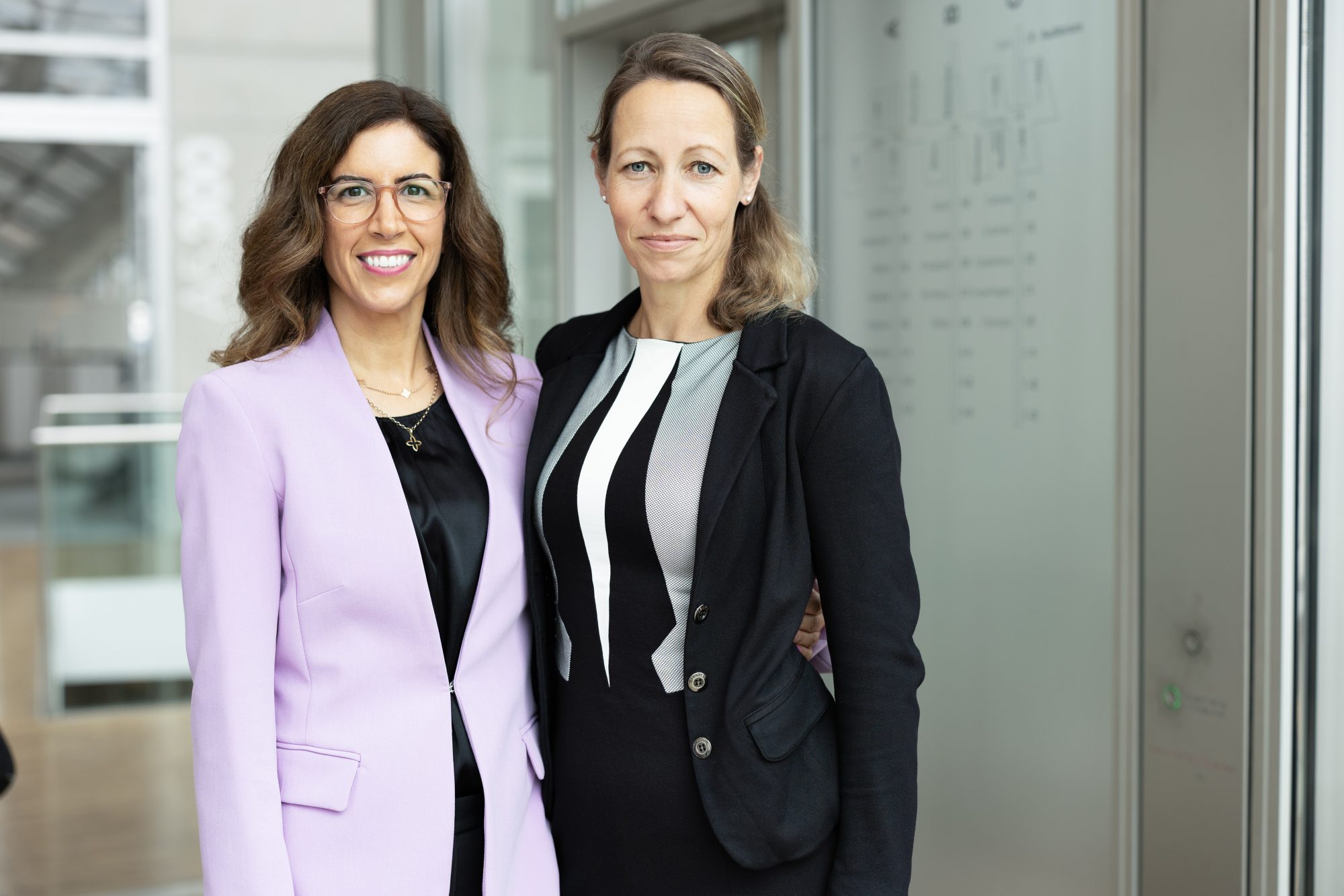 Zwei Frauen im fliederfarbenen und schwarzen Blazer vor einer Glasfassade.