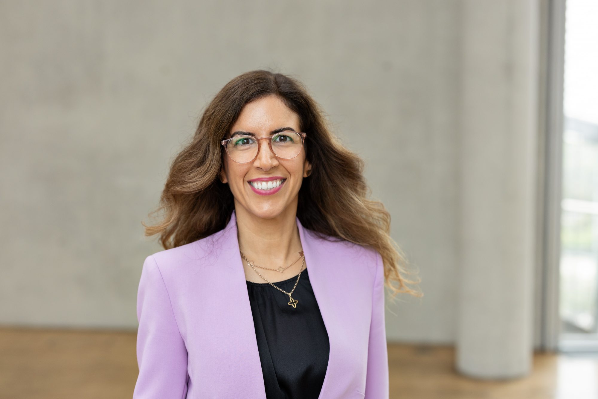 Lächelnde Frau mit Brille und fliederfarbenem Blazer vor einer Betonwand.