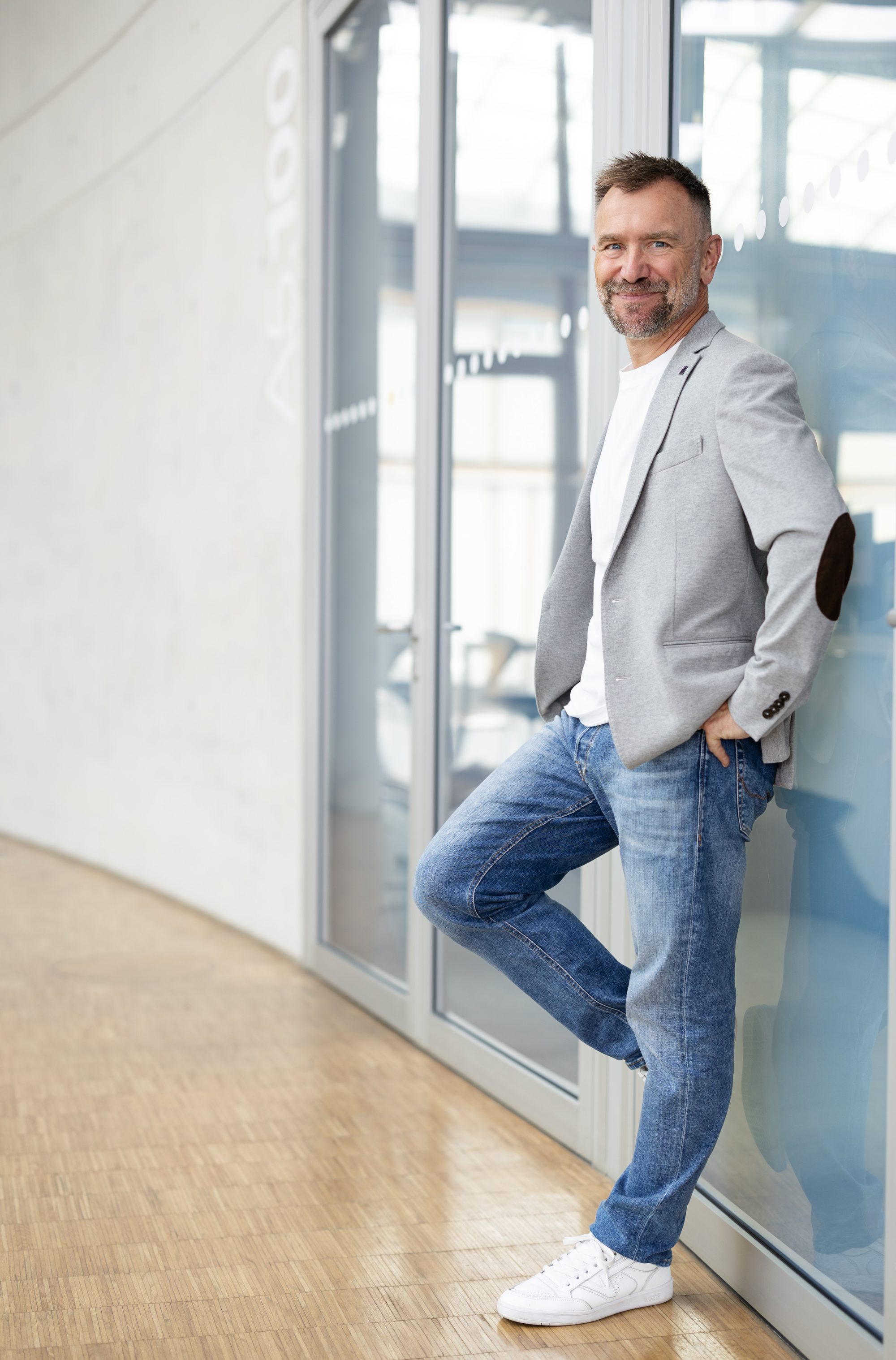 Mann im grauen Sakko und Jeans steht entspannt an einer Fensterfront.