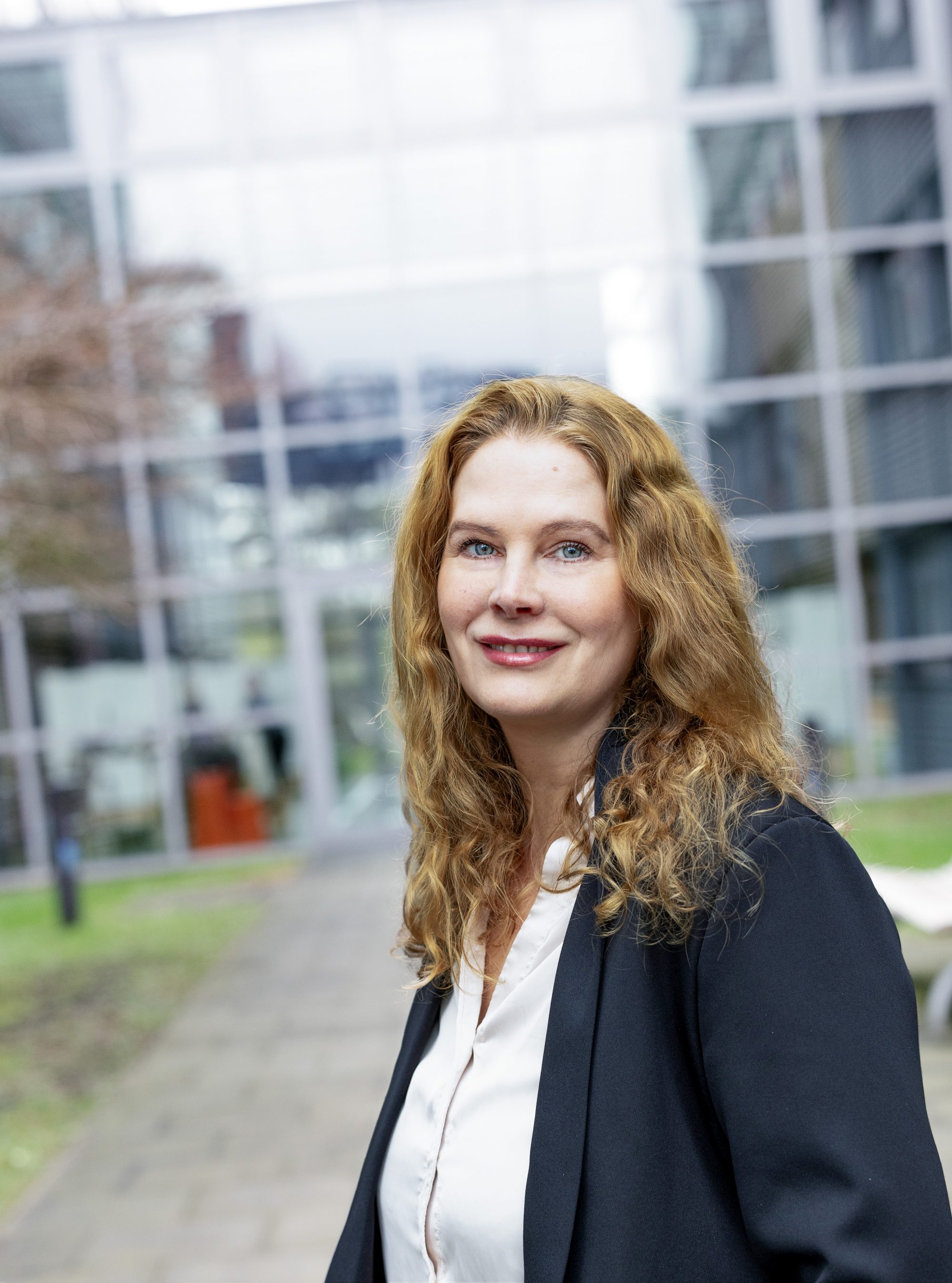 Frau im dunklen Blazer steht lächelnd im Außenbereich vor einer Glasfassade.