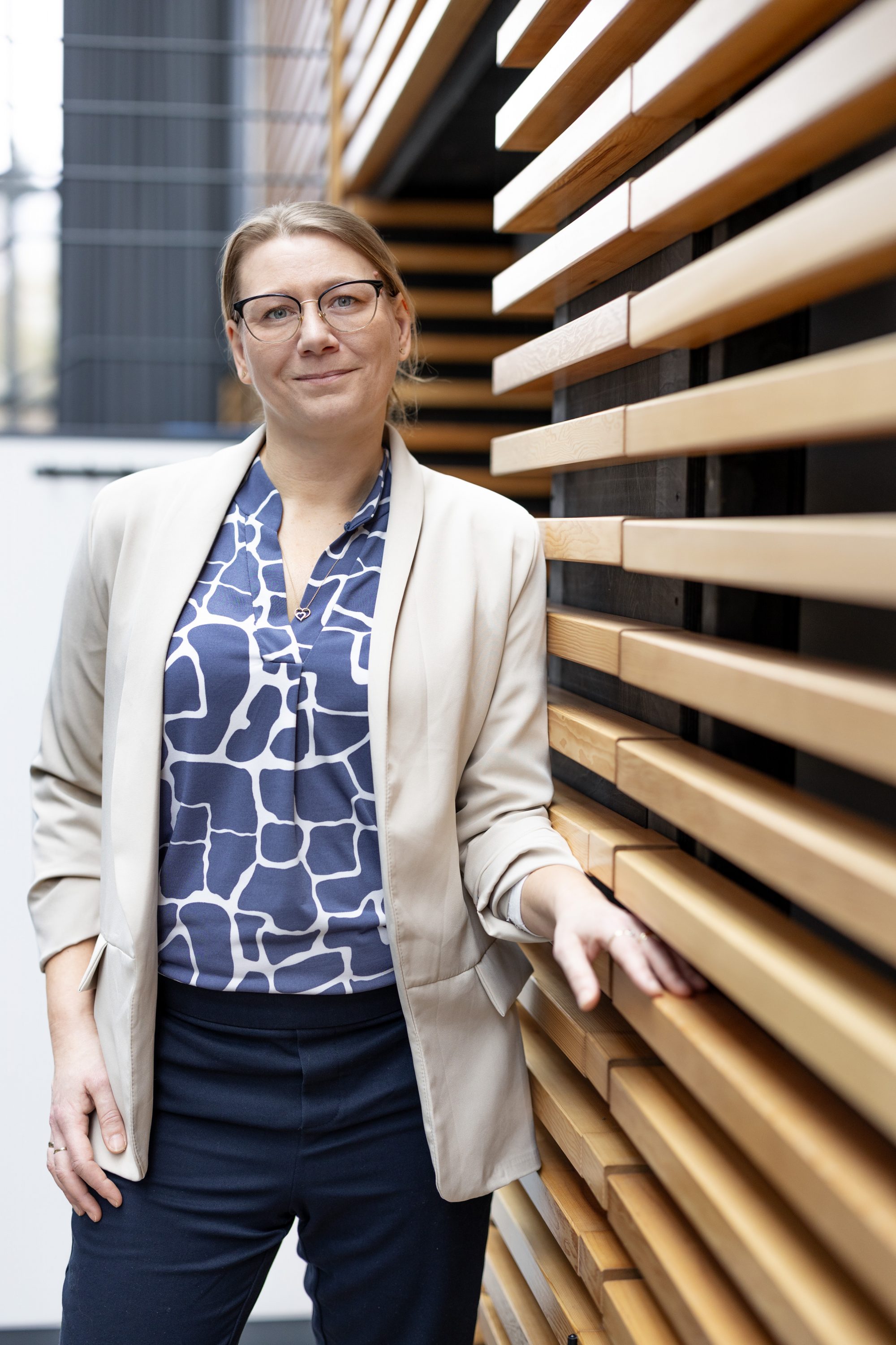 Frau mit Brille und hellem Blazer steht neben einer Wand aus Holzlamellen.