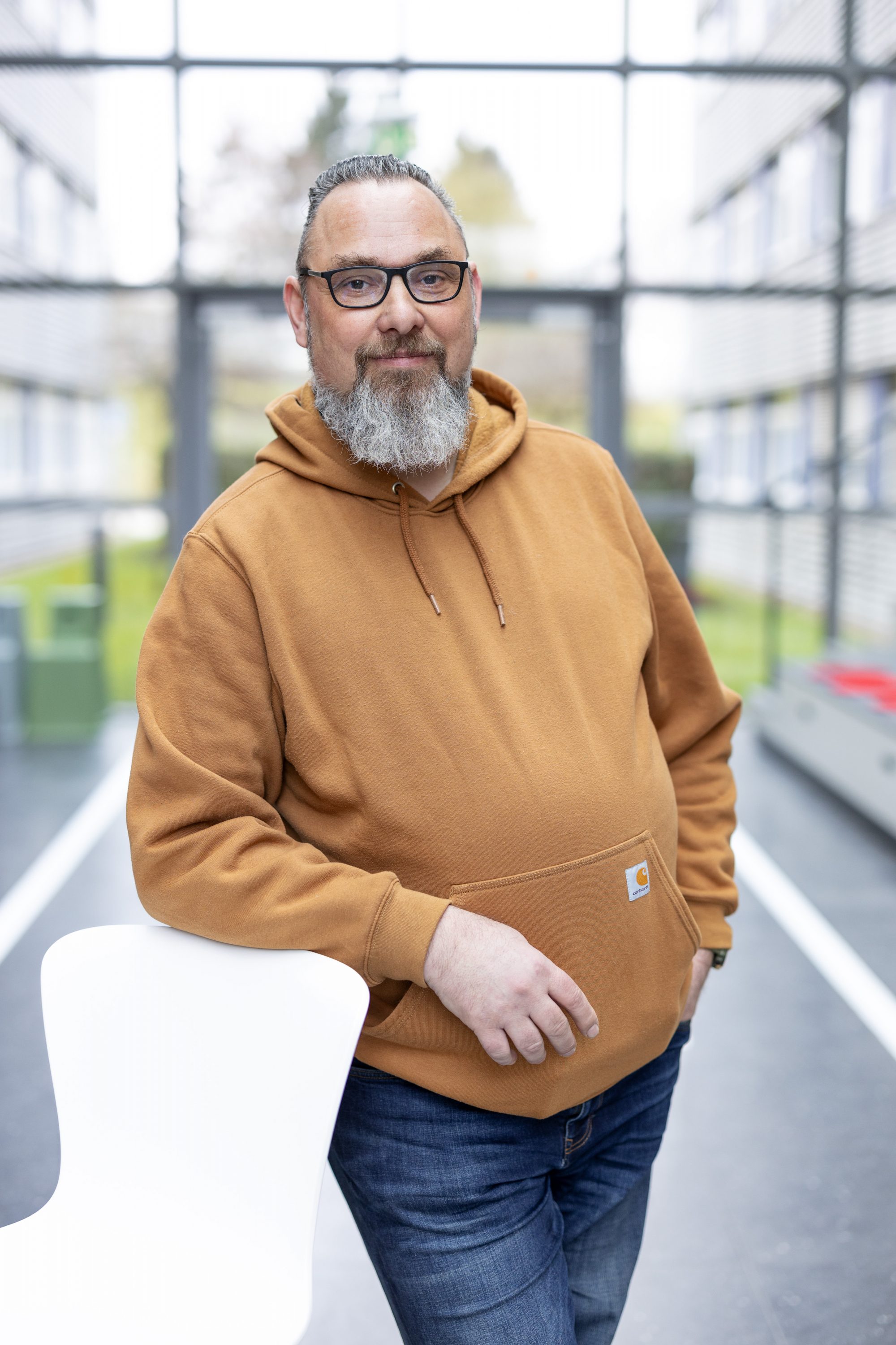 Mann mit Brille und Bart im ockerfarbenen Pullover in einem verglasten Gebäude.