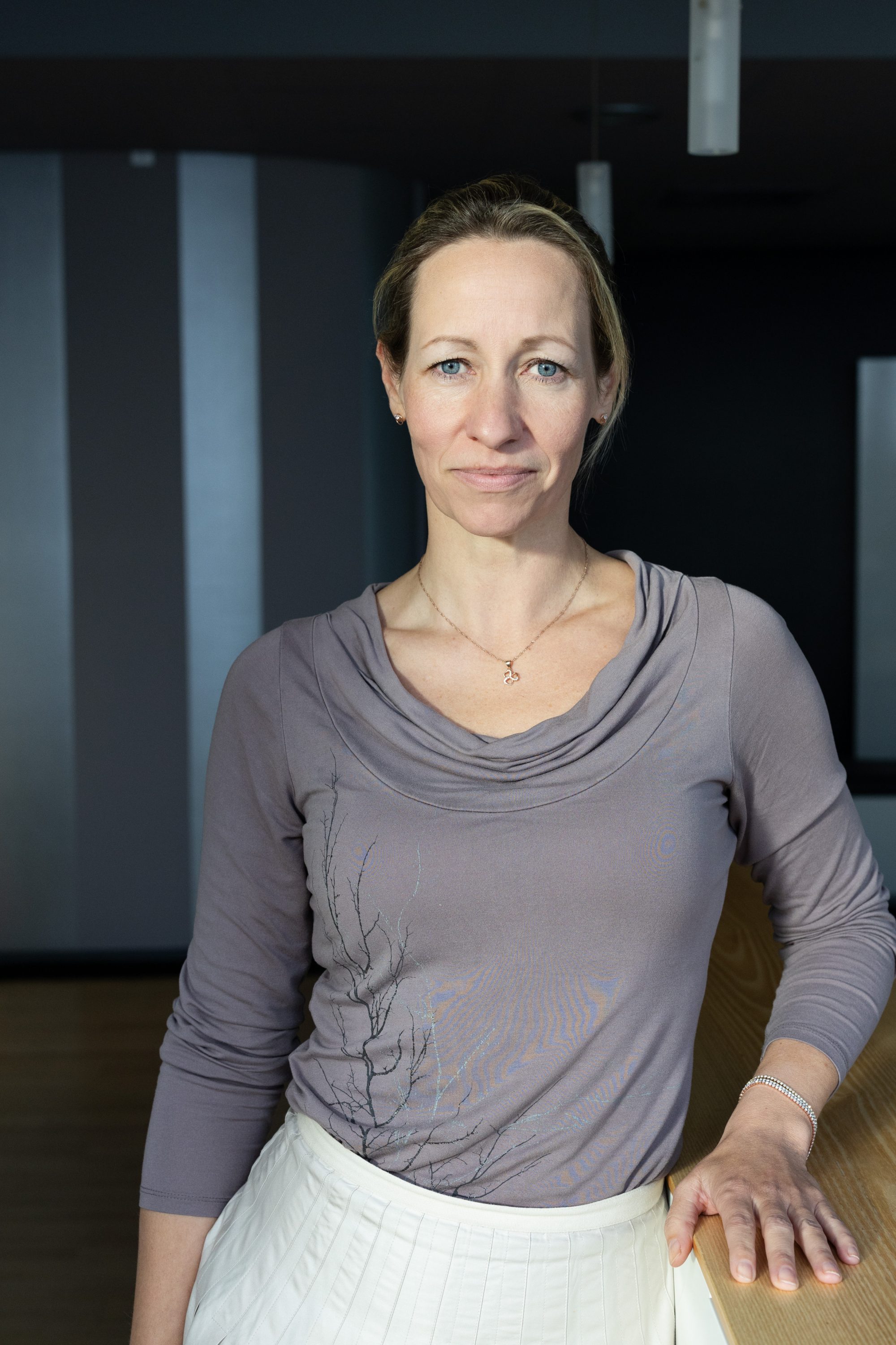 Frau im grauen Oberteil steht neben einer Holzfläche vor einem dunklen Hintergrund.