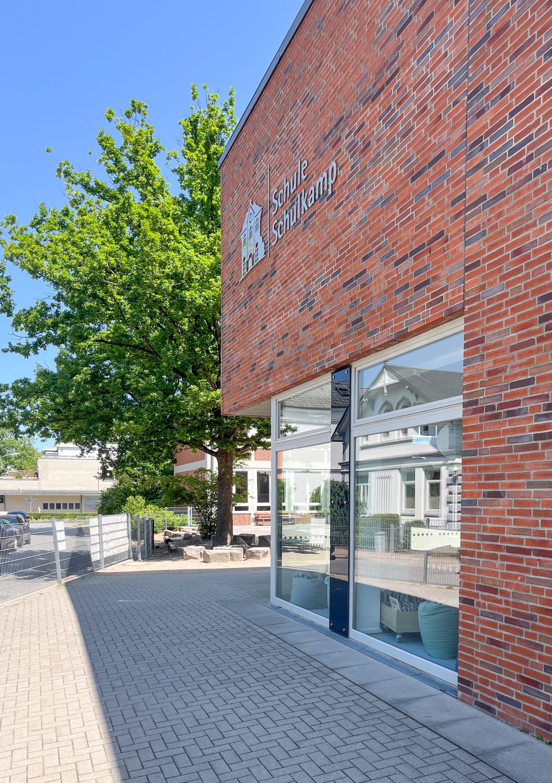 Backsteinfassade mit breiter Glasfront und gepflastertem Weg auf dem Schulgelände.