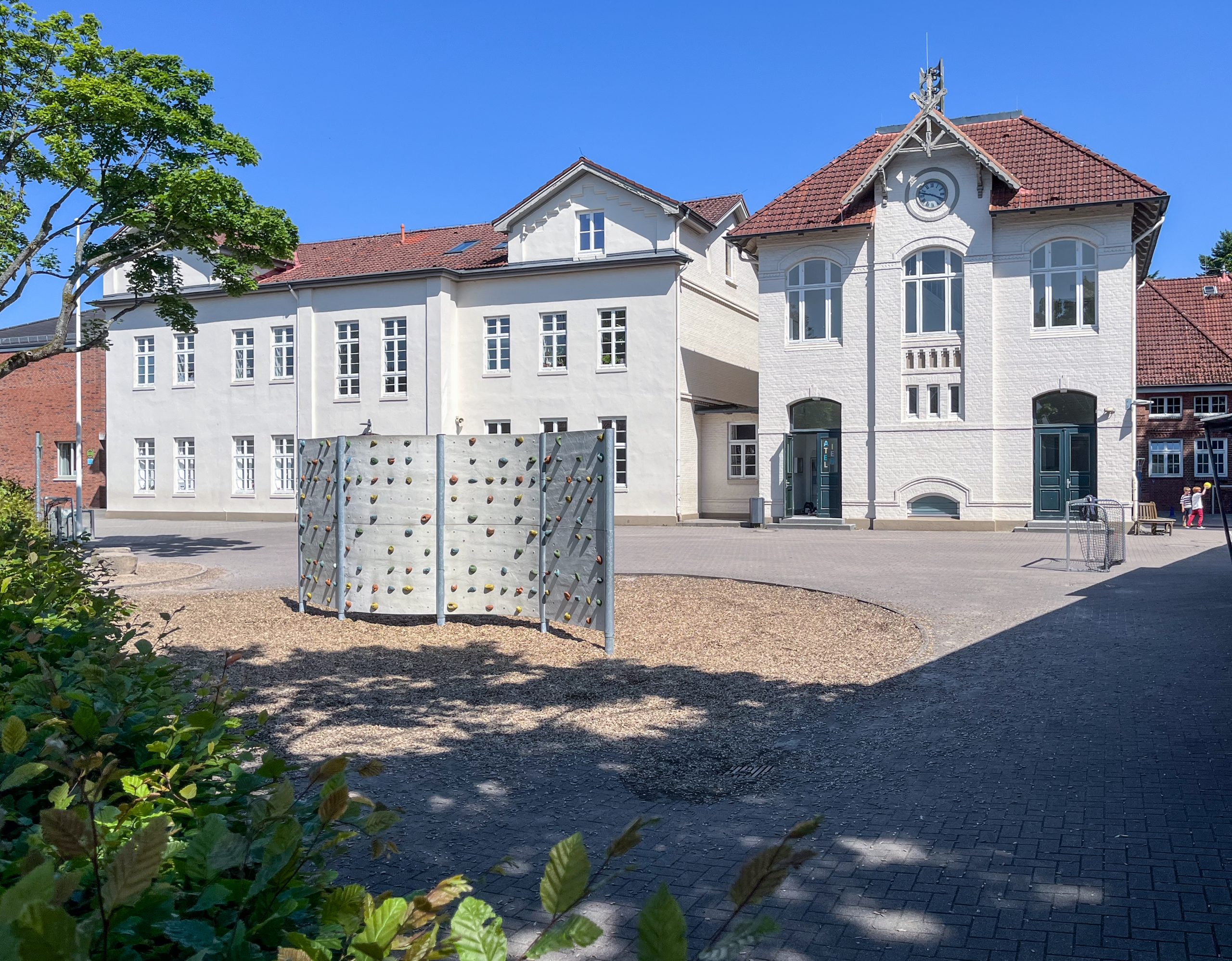 Helles Schulgebäude mit Uhrgiebel hinter dem Schulhof und einer Kletterwand.