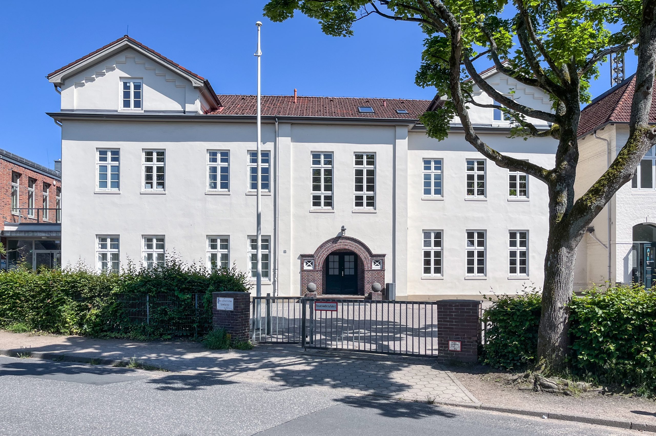 Straßenansicht des hellen Schulgebäudes mit Uhrgiebel, Eingangstor und Baum.