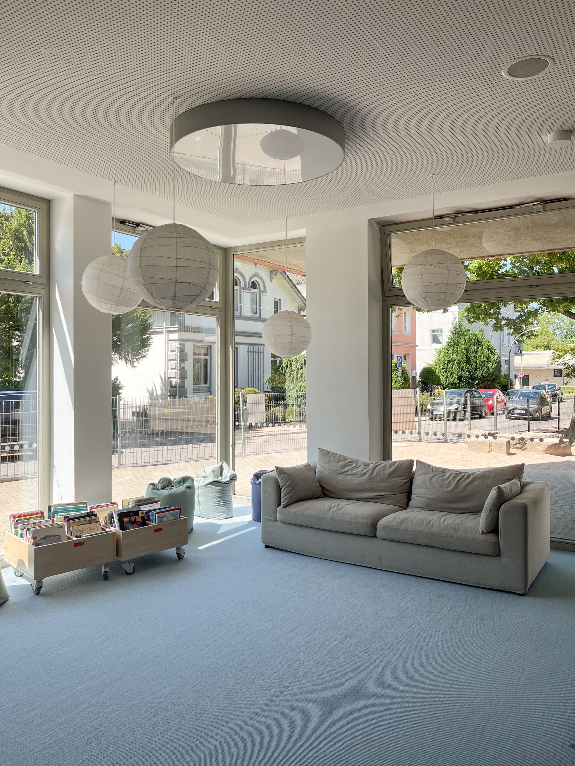 Heller Aufenthaltsraum mit grauem Sofa und großen Fenstern.