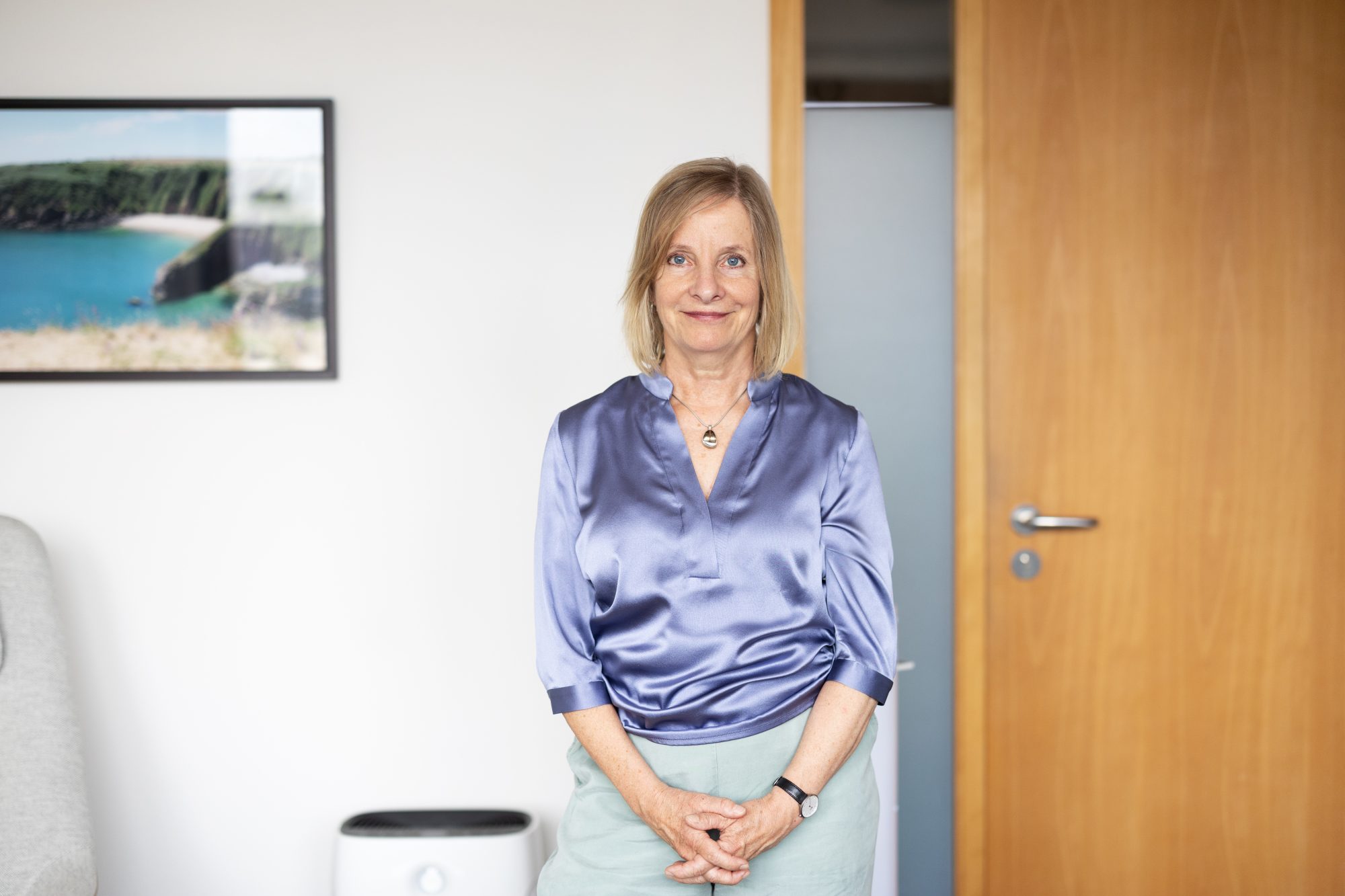 Nanette Toillié sitzt in einer blau schimmernden Bluse vor einer Holztür in der Praxis.