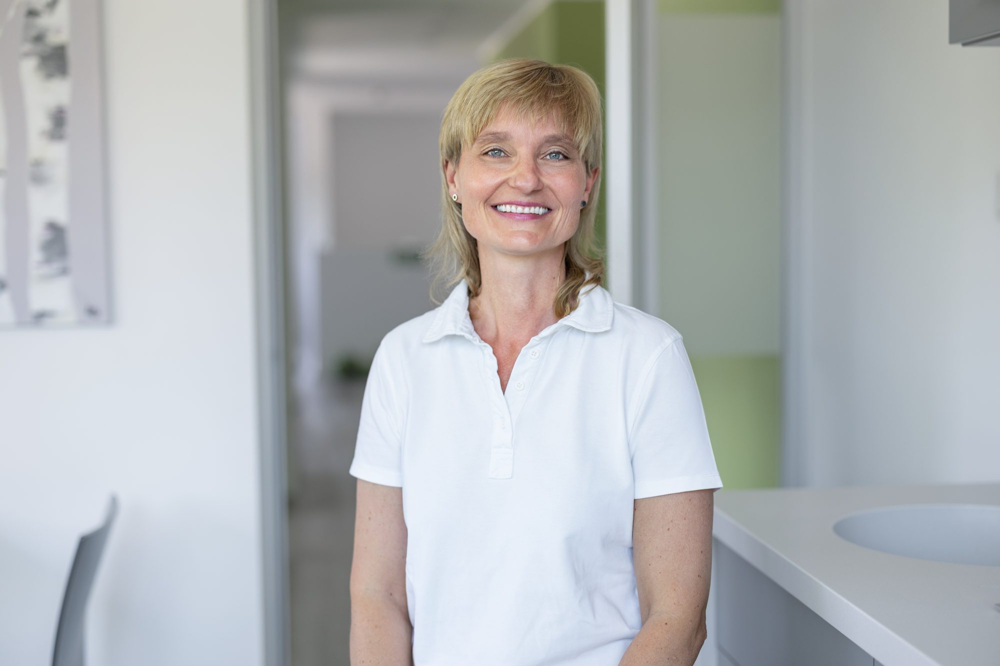 Lächelnde Frau im weißen Praxisoberteil sitzt an einer Arbeitsfläche in der Kieferorthopädie Walddörfer.