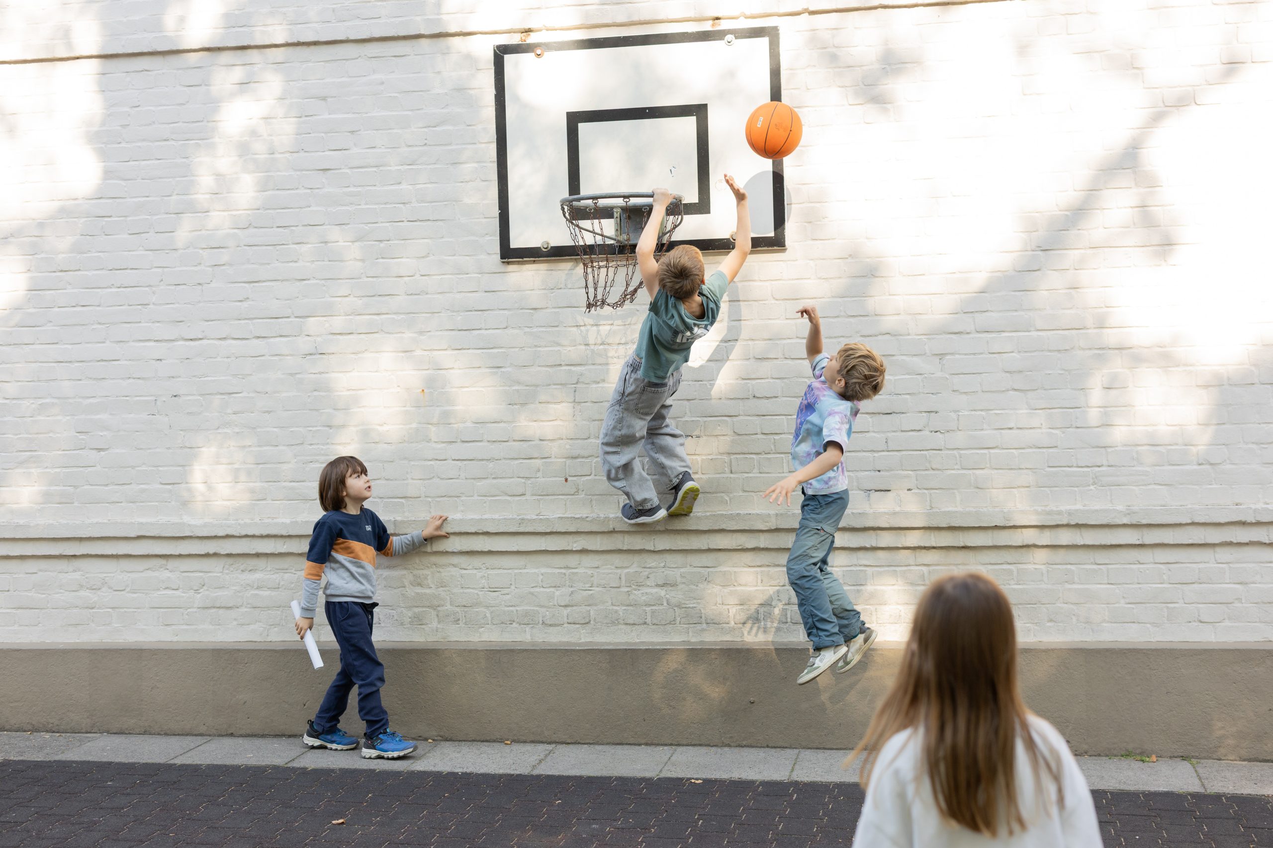 Kinder springen beim Basketballspiel vor dem Korb an einer hellen Backsteinwand.